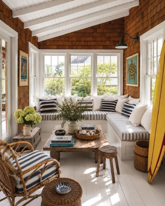 A shingled Fire Island sunroom with striped cushions and a yellow surfboard